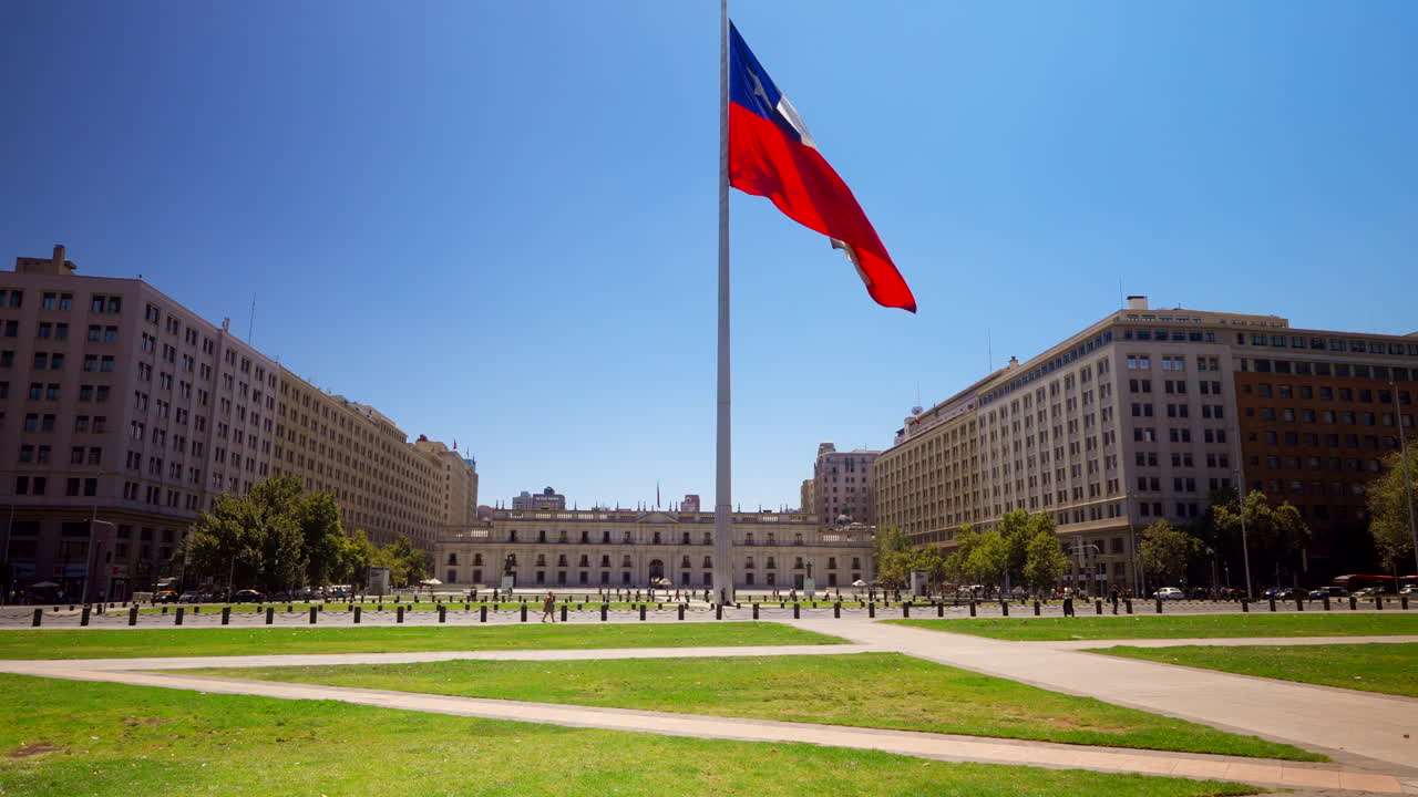 Chilean flag waves near the Centro Cultural La Moneda in downtown Santiago, Chile, towering above cars, static establishing