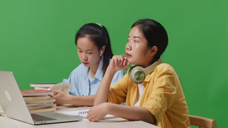 close up of asian woman student thinking and looking around while sitting with her friend and writing into the book on table with a laptop in the green screen background classroom