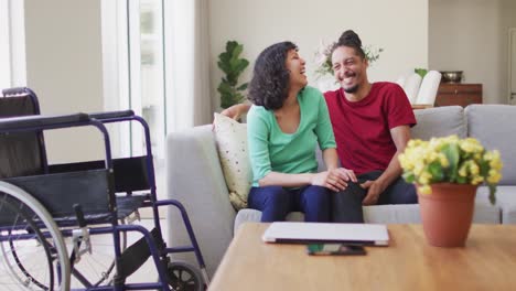 Happy-biracial-couple-sitting-on-couch-laughing-in-living-room,-with-wheelchair-in-foreground