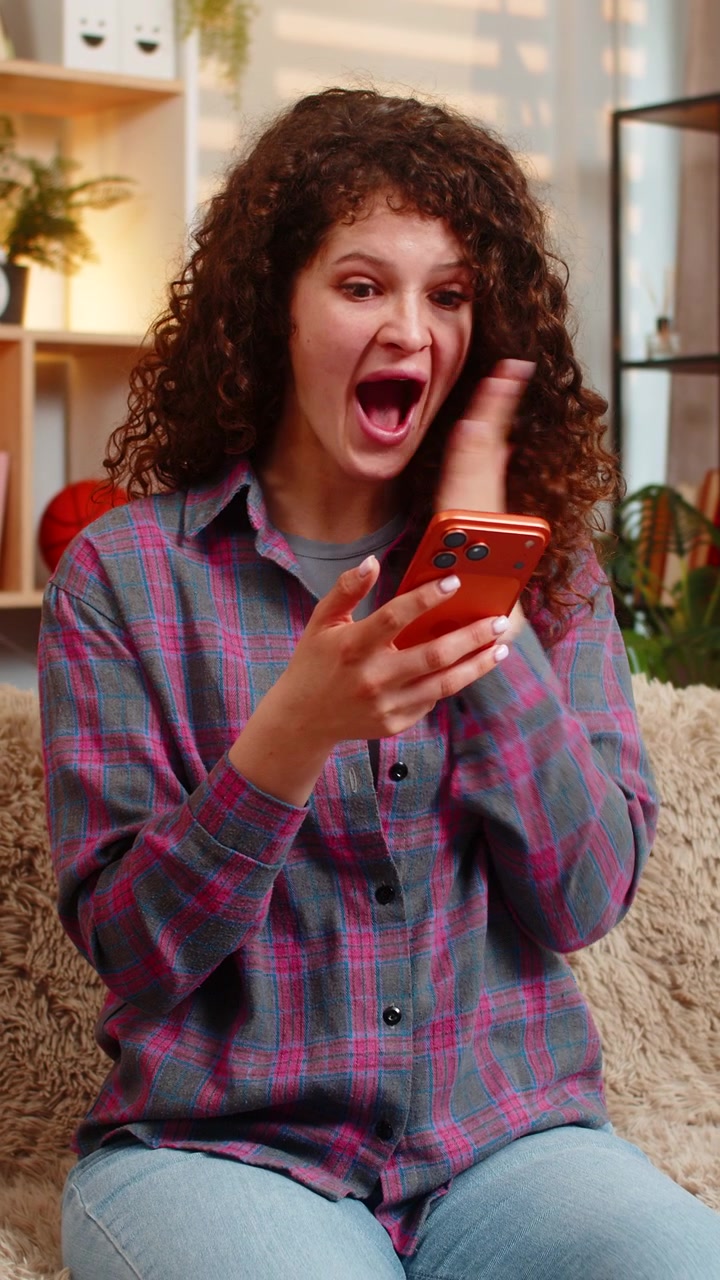 Giovane donna sul divano di casa reagisce allo smartphone con un sorriso di stupore dopo un'offerta a sorpresa, un regalo o un messaggio.