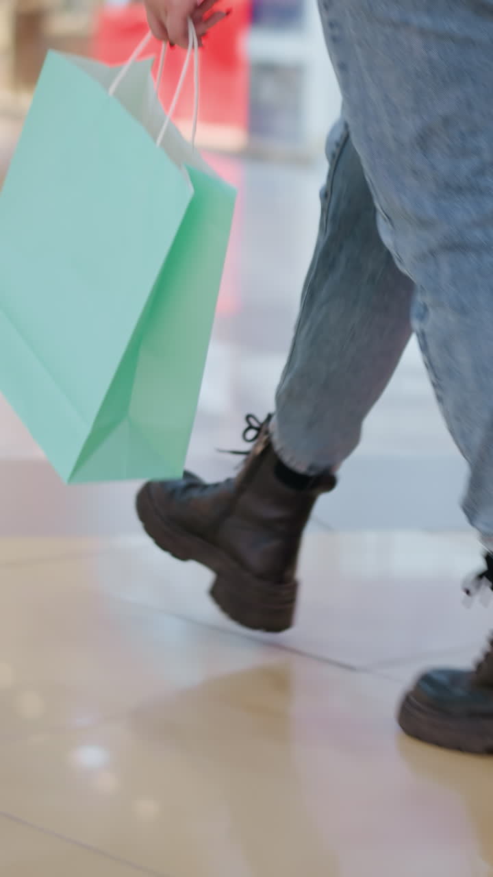 vista lateral de la pierna de una persona con botas negras y vaqueros caminando a través de un centro comercial moderno mientras lleva bolsas de compras, el piso de azulejos elegante y el fondo borroso con otros compradores