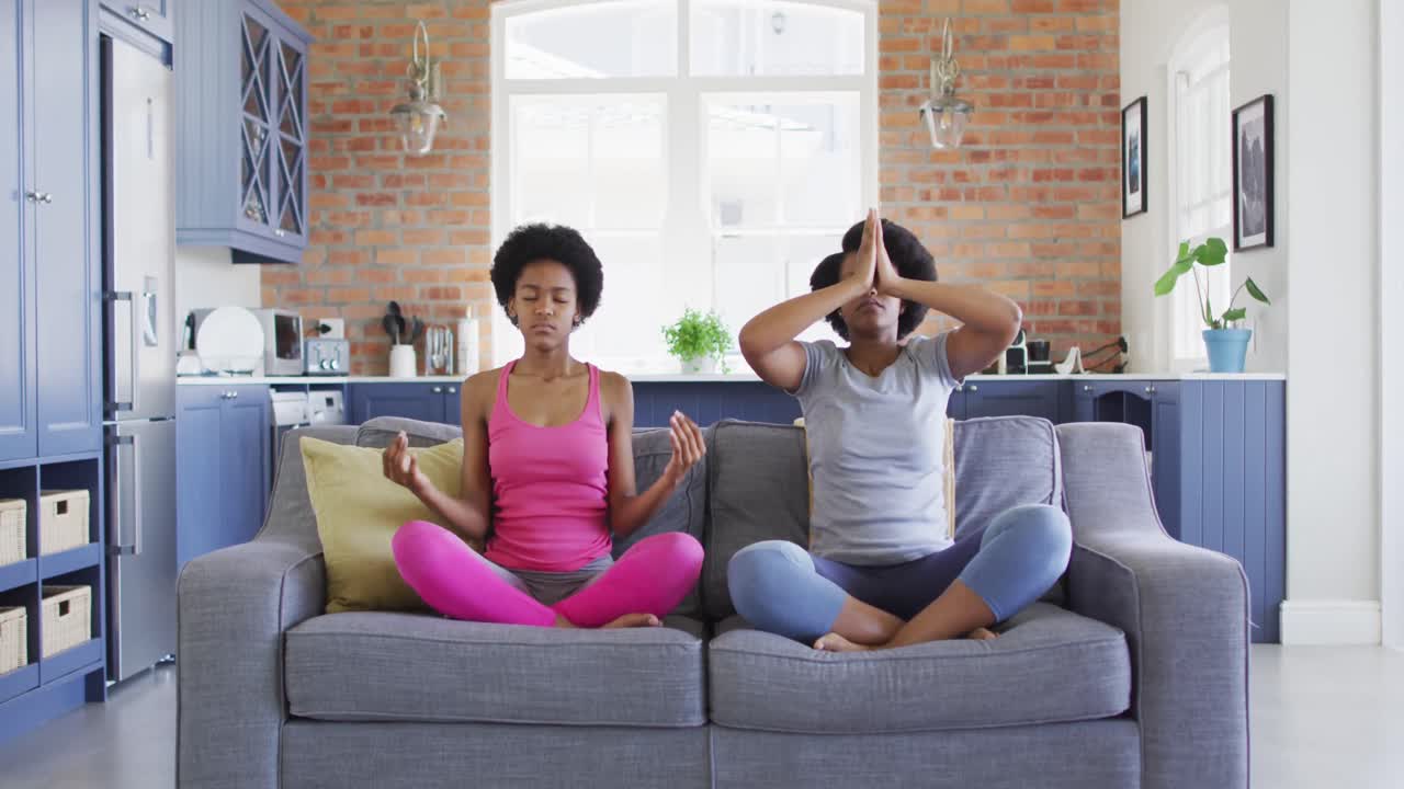 Relaxed african american mother and daughter doing yoga in living room, meditating