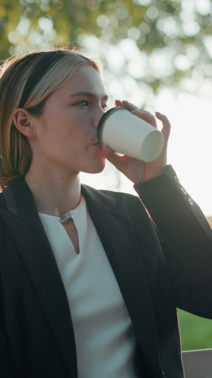 Elegant remote worker focuses on laptop while sipping water from cup, seated on bench in sunny outdoor park with trees, distant moving bus, parked cars, and urban building