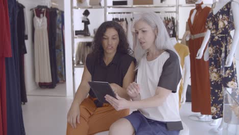 two women sitting together in fashion store with tablet, talking