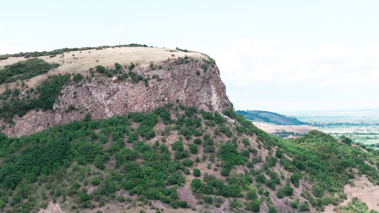 Aerial tele drone shot orbits the Măgura Uroiului mountain in Romania. The view showcases the sheer rock cliff, green slopes, and the vast Transylvanian plain below