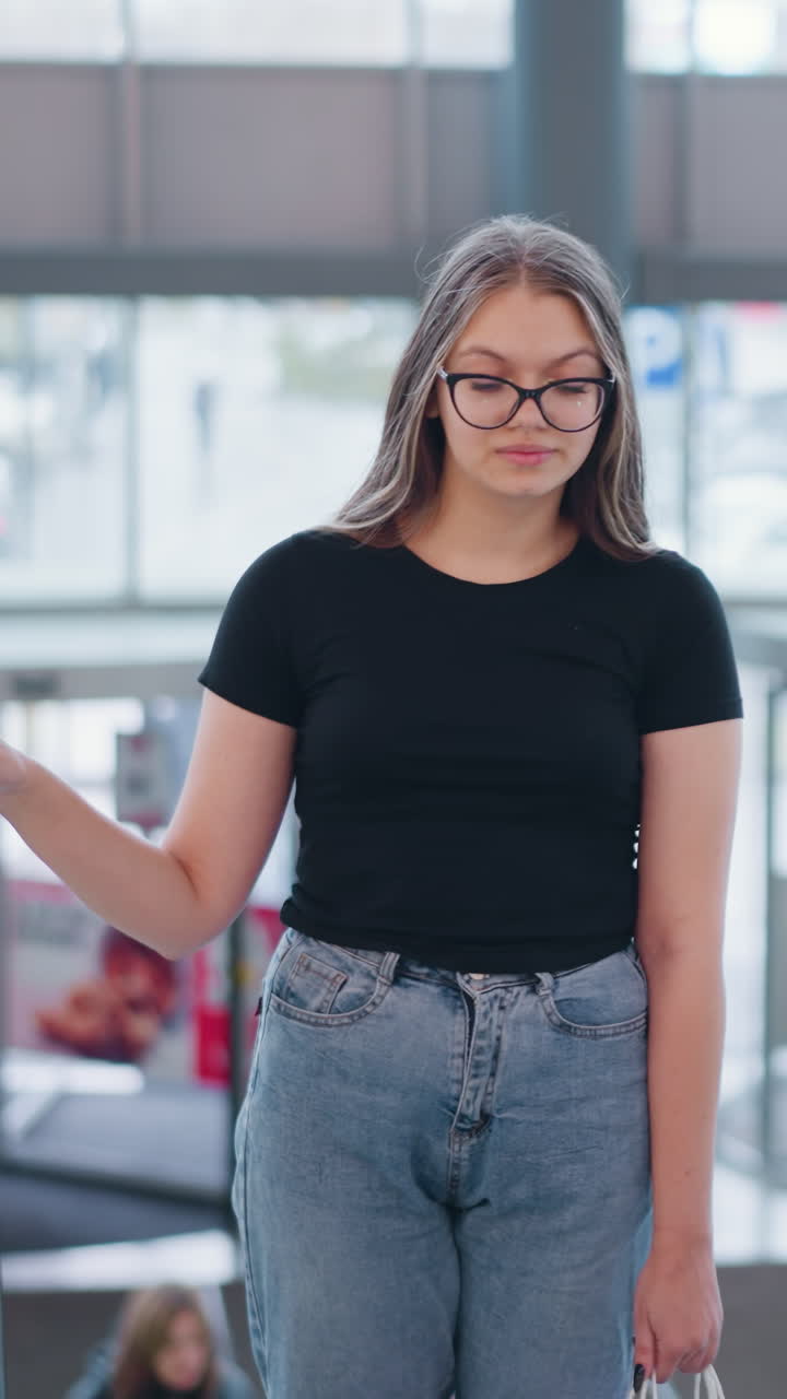 una mujer joven con una camiseta negra y pantalones vaqueros sube por la escalera mecánica en el centro comercial sosteniendo dos bolsas de compras, con otros compradores borrosos en el fondo, reflejando una escena urbana cotidiana