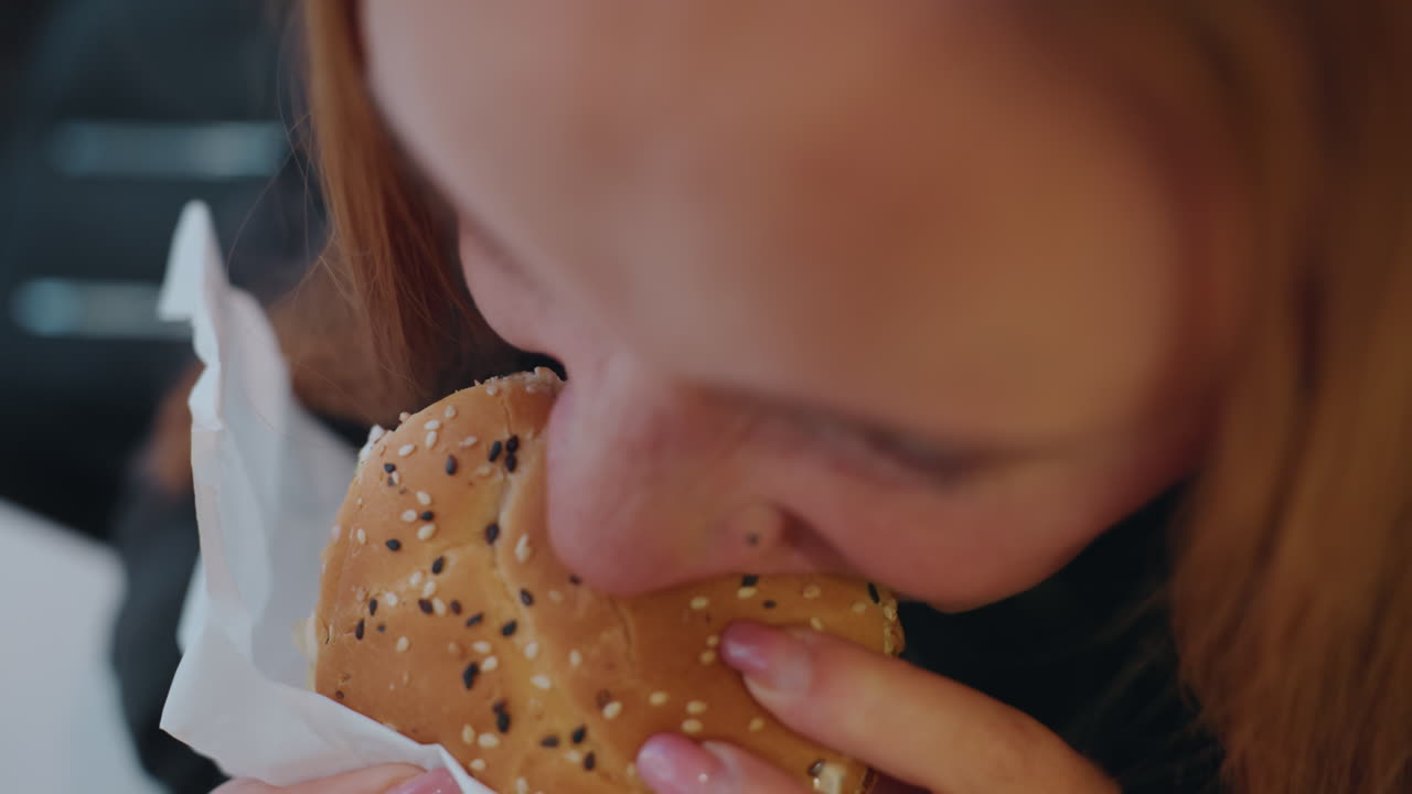 Young lady bites sesame sandwich. Freckled redhaired woman relaxes and eats her sandwich contentedly. Relaxed young woman with red hair and freckles enjoys sesame burger quietly in cozy cafe
