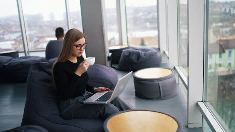 beautiful lady in office near the window. attractive young business woman working on a laptop and drinking coffee. pretty girl in comfortable place having break during work.