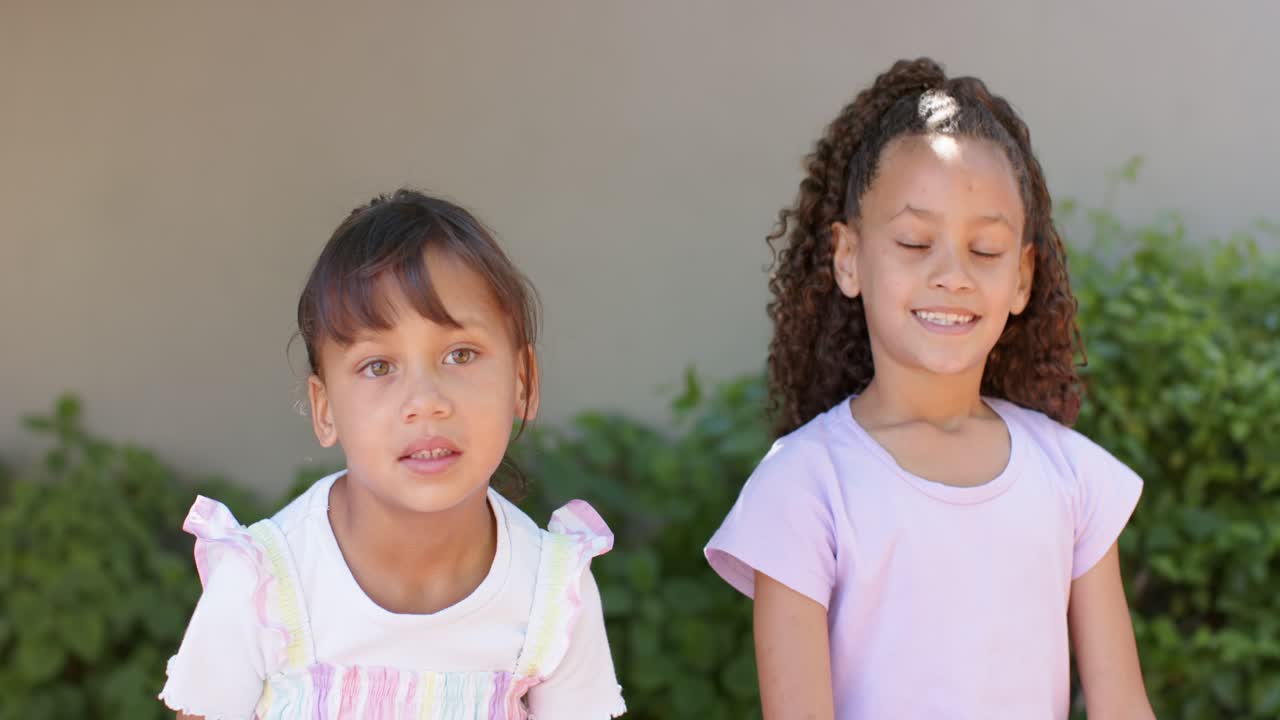 Ragazze preadolescenti afroamericane in posa per la macchina fotografica vicino a un muro beige e arbusti, con un vestito color pastello e una maglietta viola.