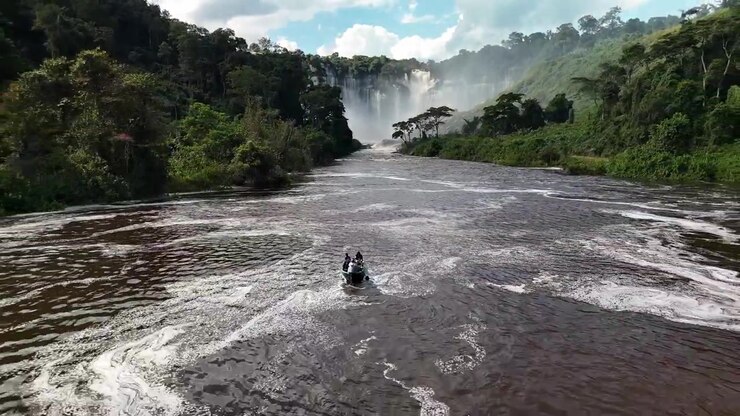 Panoramica suggestiva di una piccola imbarcazione su un fiume a scorrimento rapido con la nebbia nebulizzata delle cascate di Kalandula in lontananza, circondata da una fitta foresta tropicale nella provincia di Malanje, Angola