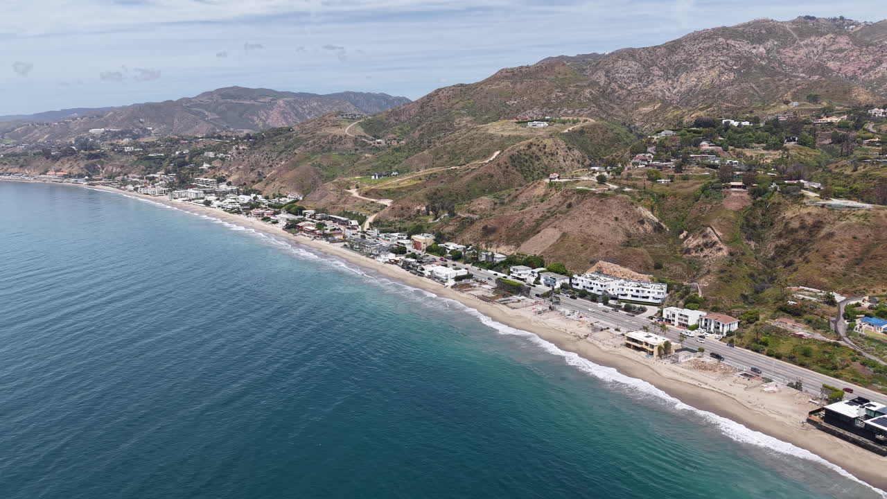 Riprese aeree cinematografiche diurne con drone catturate in alto sopra l'oceano al largo della costa di Malibu, con un movimento di discesa fluido combinato con un carrello in avanti verso la costa