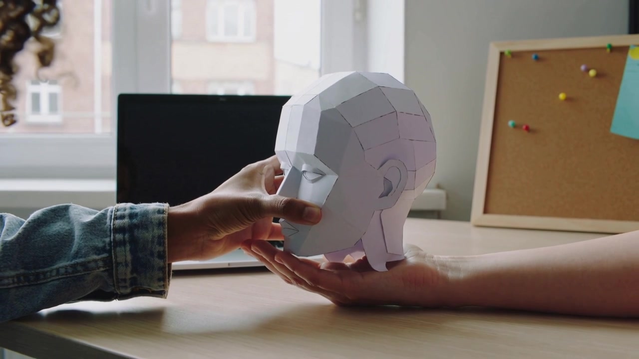 Lowering left hand wearing denim jacket guiding papercraft head onto right palm at desk for display