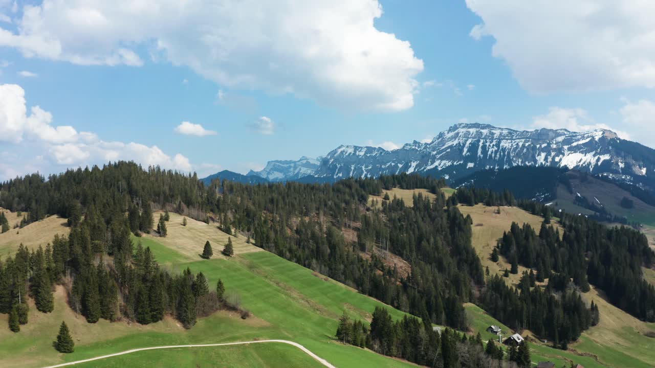 Jevnt luftfoto av fjellandskap i Entlebuch, Sveits, om våren, med furutrær og snødekte fjell under en skyet himmel.