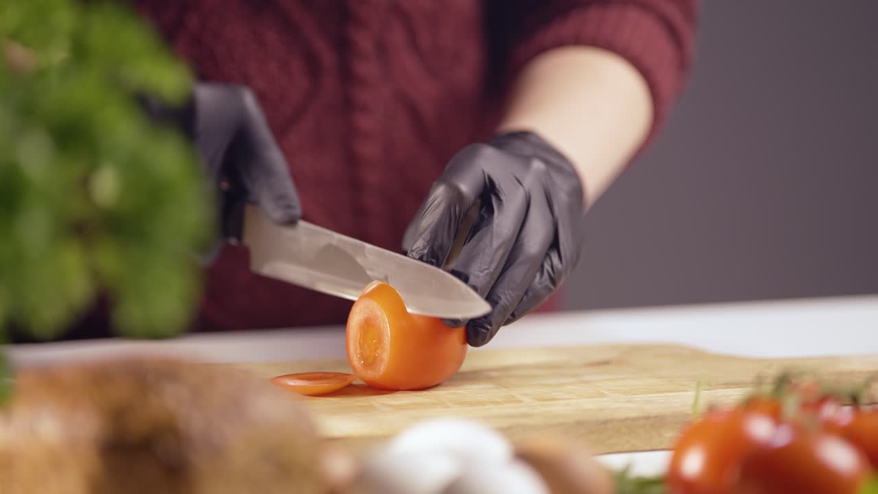 Slow-motion close-up of a chef slicing a ripe tomato on a wooden board. Black gloves ensure hygiene, while a sharp knife glides smoothly. Fresh ingredients like herbs, bread, and eggs add depth