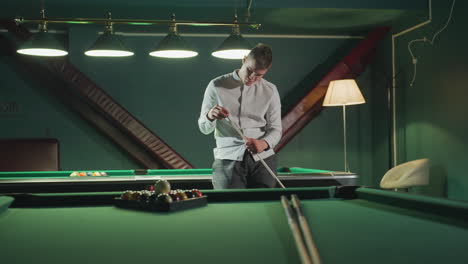 sportsman in white shirt and grey trousers sits on green pool table, holding cue stick with focused expression. another pool table with arranged billiard balls is visible in background