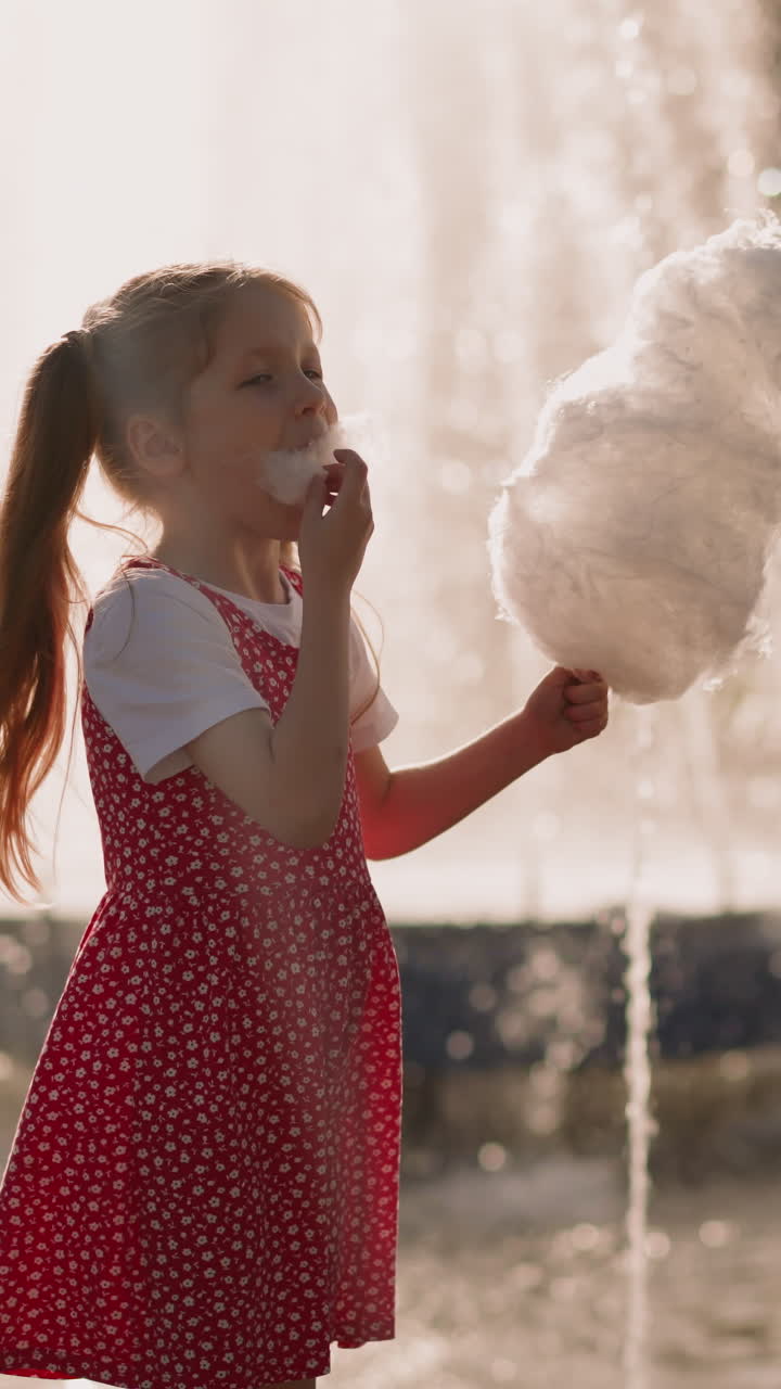 Preschooler girl with younger brother eats cotton candy standing near fountain on square slow motion. Carefree children spend time in amusement park
