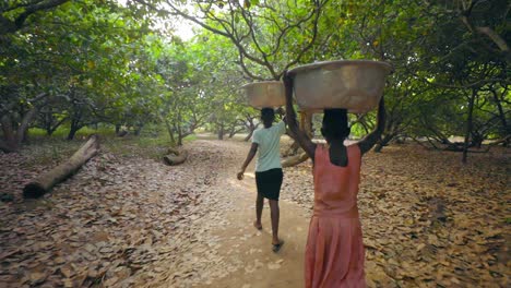 Group-of-local-young-African-women-and-kids-are-carrying-big-heavy-buckets-of-drinking-water-and-bringing-the-water-on-their-heads-towards-the-village-walking-in-line-in-bush-forest-jungle---Wide-shot