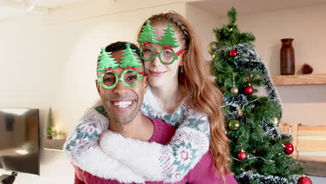 joyful couple wearing festive glasses embracing by christmas tree at home