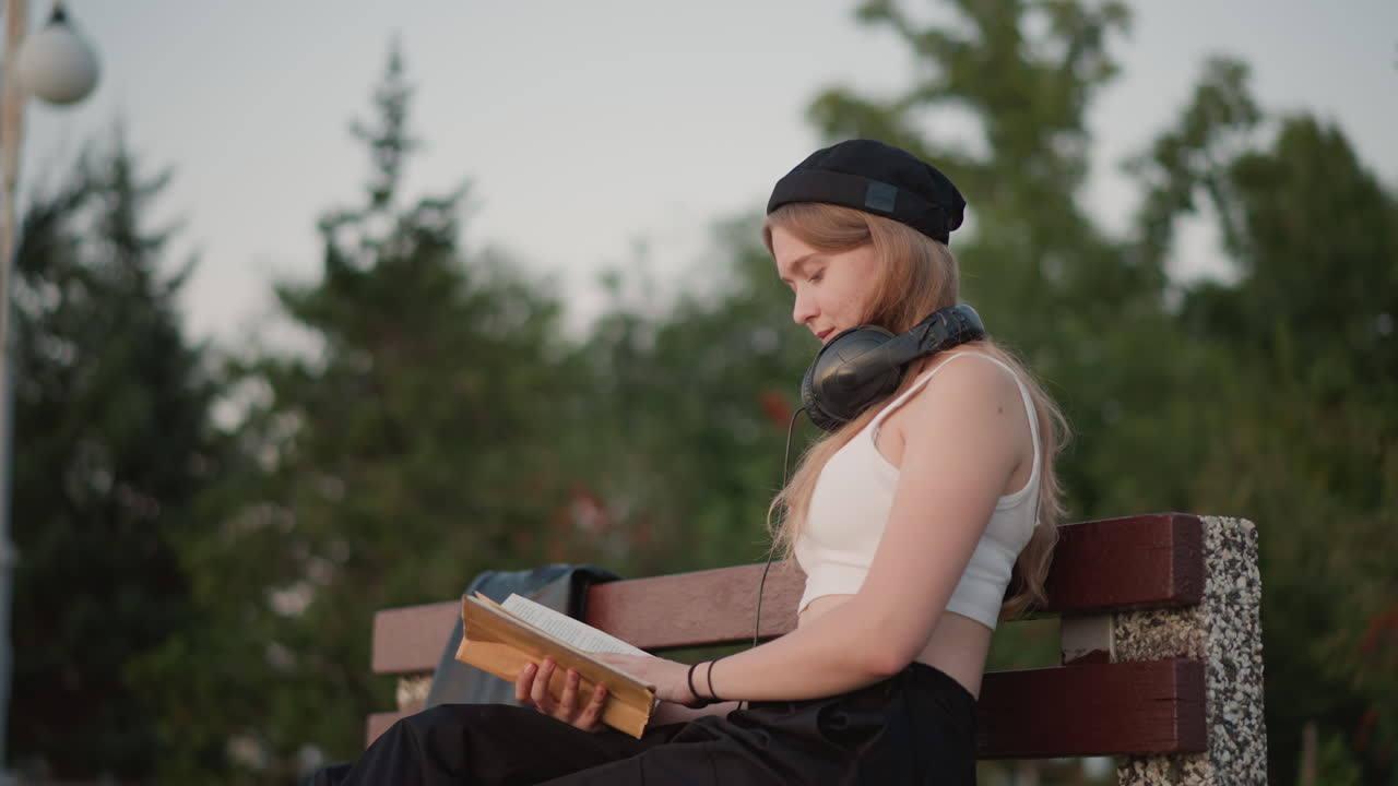 Biała kobieta czyta na ławce w otoczeniu drzew o zmierzchu, słuchawki na szyi, a beret przechylony. Pochłonięta tekstem na tle miękkiego nieba, spokojna zieleń i latarnia uliczna otaczają kameralny, refleksyjny moment