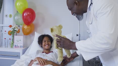 Diverse-male-doctor-playing-teddy-bear-with-girl-in-hospital-bed-wearing-fingertip-pulse-oximeter