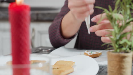 close-up of red burning candle with tray of cookies and decorative flower as woman uses piping bag to apply intricate icing details on cookie