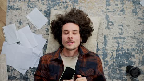 un cansado chico moreno con cabello rizado y bigote yace en el suelo y trata de descansar en una almohada en casa en un apartamento moderno