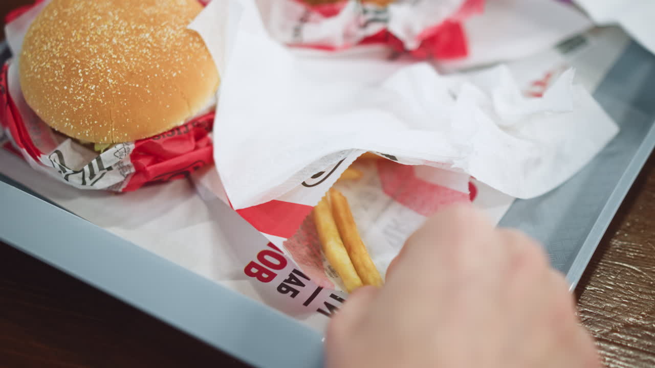 Close up view of KFC fries bags with hand reaching, golden crispy fries spilling from red containers, oil sheen visible, casual snack moment in fast food setting, hunger and craving evident