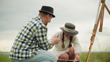 a cheerful moment captured as an artisan wearing a hat and checked shirt struggles to open a paint tube. a woman in a white dress and hat, smiling warmly, assists by taking the tube to help him