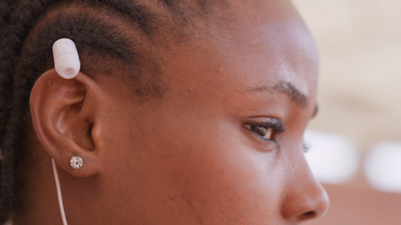 Profilo di ragazza nera che ascolta in classe, capelli intrecciati con auricolare in schiuma bianca, orecchino a bottone con diamante, luce naturale soffusa, primo piano di profilo che mostra espressione concentrata e sottile emozione, stato d'animo.