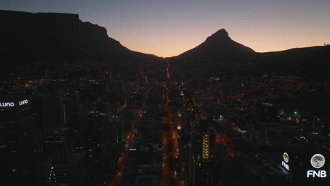 Volar-Sobre-La-Ciudad-De-Noche.-Silueta-De-Montañas-Contra-El-Pintoresco-Cielo-Crepuscular.-Al-Revés-Revelando-Los-Edificios-De-Oficinas-Del-Centro-De-Gran-Altura.-Ciudad-Del-Cabo,-Sudáfrica