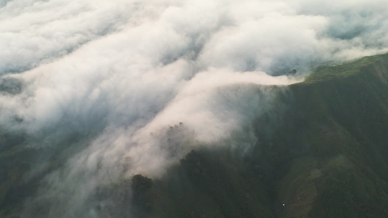 Dramatic aerial close-up of thick, fast-moving clouds or fog aggressively rolling and spilling over a steep, sharp mountain ridge. Captures the dynamic movement and sheer scale of nature. High-value