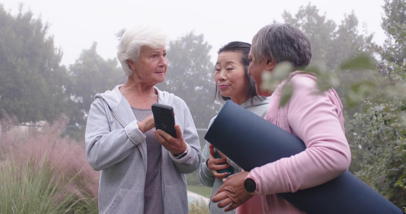Anziano con due donne che arrivano al parco; una donna vestita di rosa tiene un tappetino arrotolato e usa il telefono per organizzare lo yoga