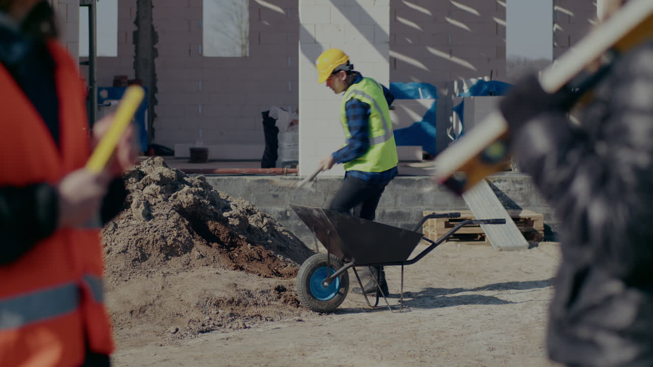 Young male construction worker collecting sand in wheelbarrow with shovel while female coworkers discussing at site on sunny day