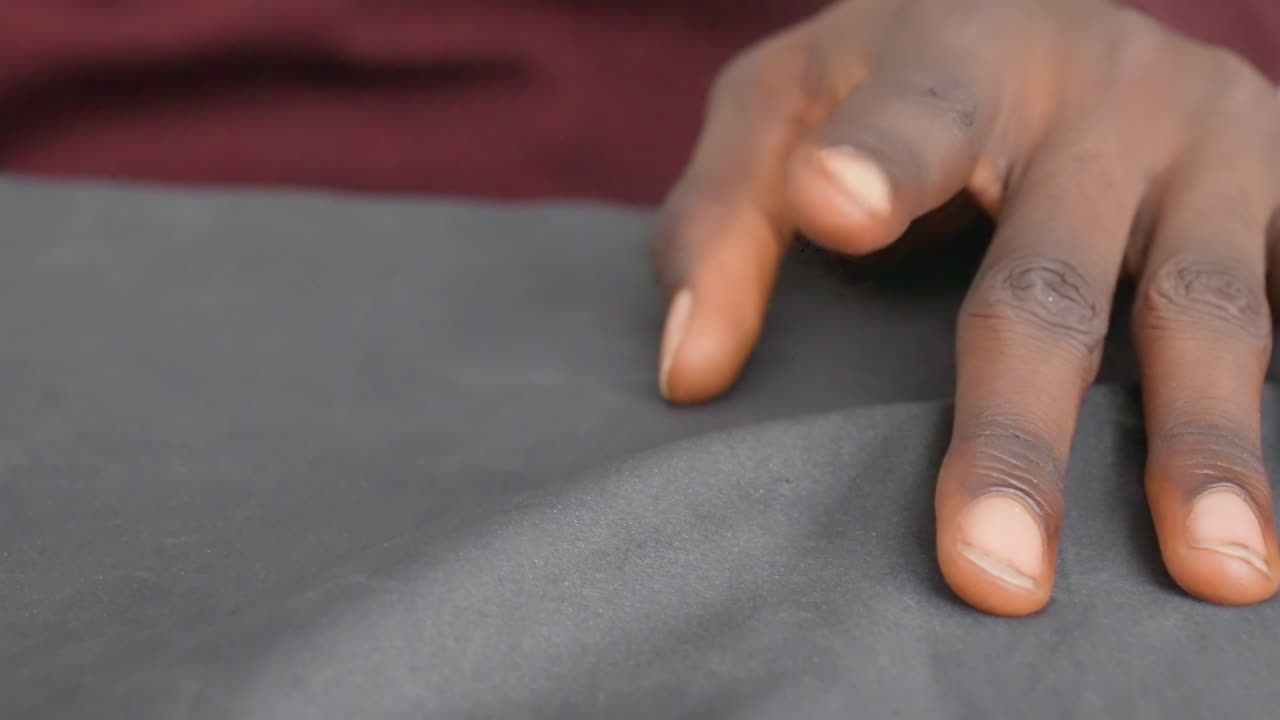 Tangan berkulit hitam di atas kanvas lukisan hitam, close-up intim jari-jari seniman berkulit hitam menjelajahi permukaan bertekstur dengan latar belakang kain marun, gerakan lambat dan sengaja menampakkan pigmen, kilau, bayangan.