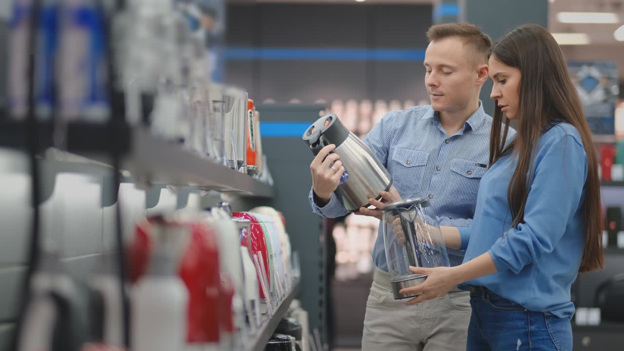 전자 제품 가게에서 캐주얼 옷을 입은 부부는 주방을 위한 전기 ketel을 선택합니다. 손에 들고 가격표와 특징을 읽고 읽습니다.