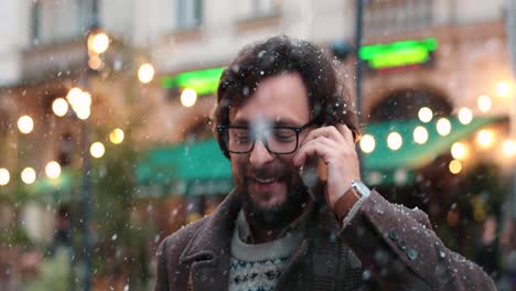 vista de cerca de un hombre caucásico con anteojos hablando por teléfono inteligente en la calle mientras nieva en navidad