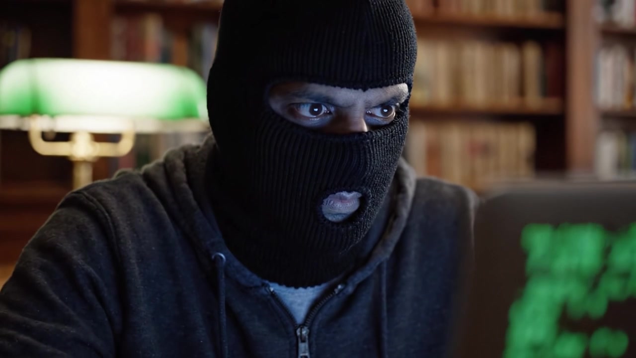 En mystisk figur i en sort skimaske udfører spændingsfyldt computeraktivitet i et dæmpet oplyst bibliotek, omgivet af rækker af bøger og en grøn lampe