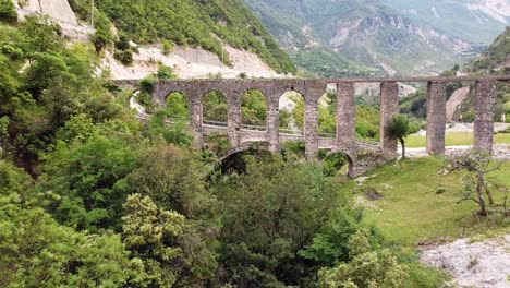 Drone-footage-of-Ali-Pasha-Aqueduct-in-Bënçë,-Albania,-capturing-the-historic-arches-surrounded-by-lush-green-hills-and-a-mountainous-landscape