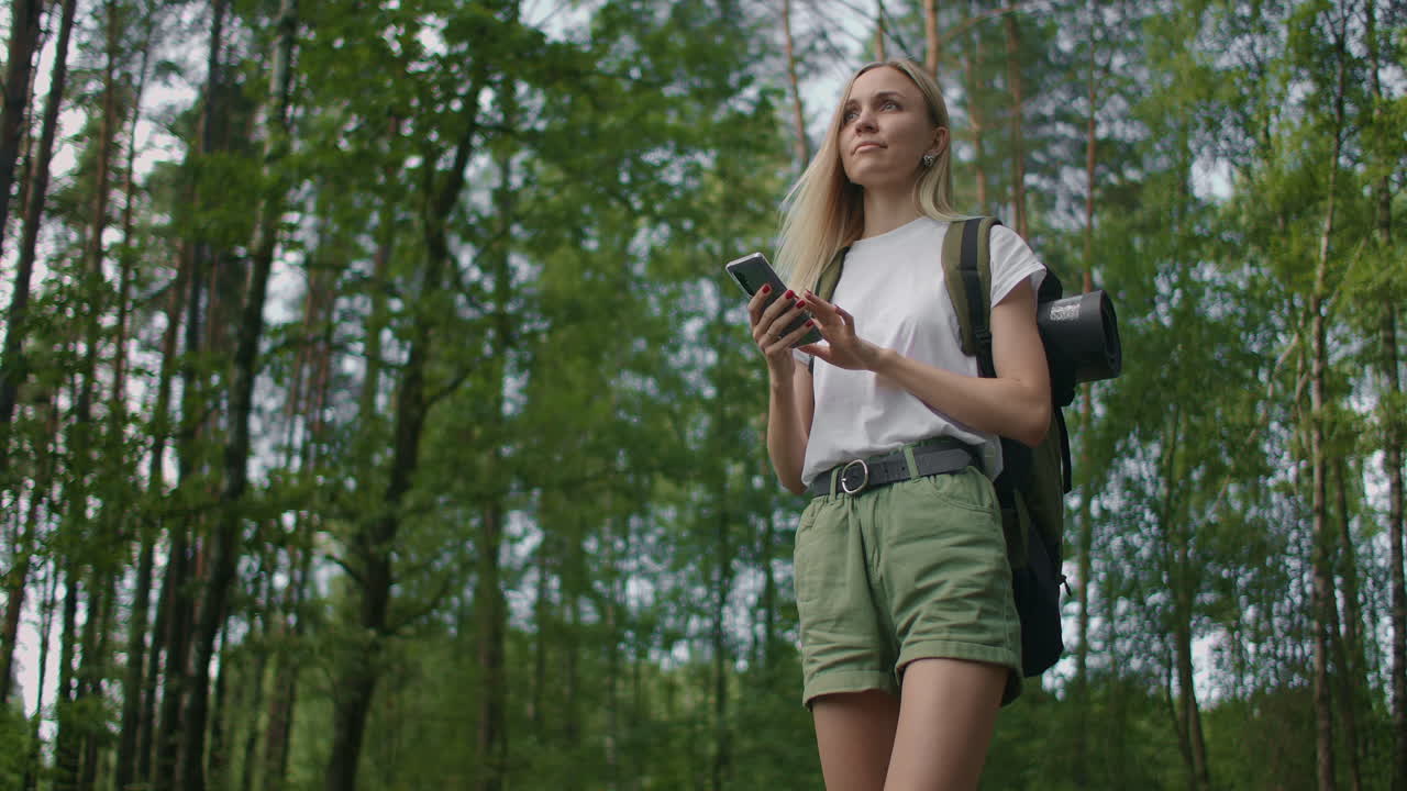 A young woman with a mobile phone walks through the forest traveling with a backpack in slow motion. Traveler in shorts in the woods looking for gps satellites