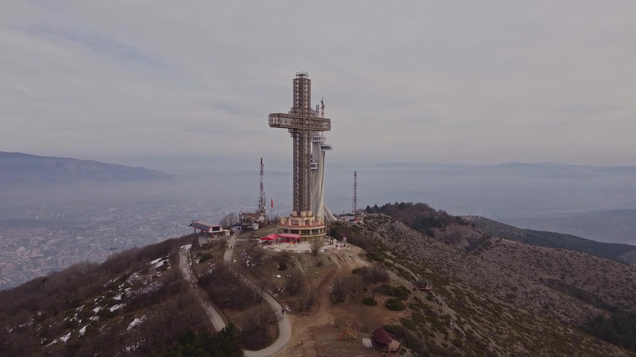 Ripresa aerea con drone in discesa e in avvicinamento alla Croce del Millennio sulla cima del Monte Vodno a Skopje, Macedonia del Nord, che mostra i sentieri per i visitatori e una vista panoramica della città nella valle nebbiosa sullo sfondo