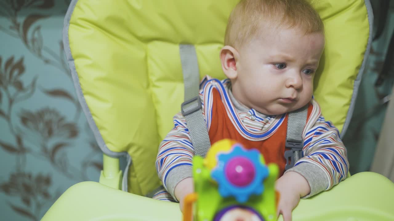밝은 플라스틱 장난감과 함께 슬픈 아기가 높은 의자 클로즈업에 앉아 있습니다
