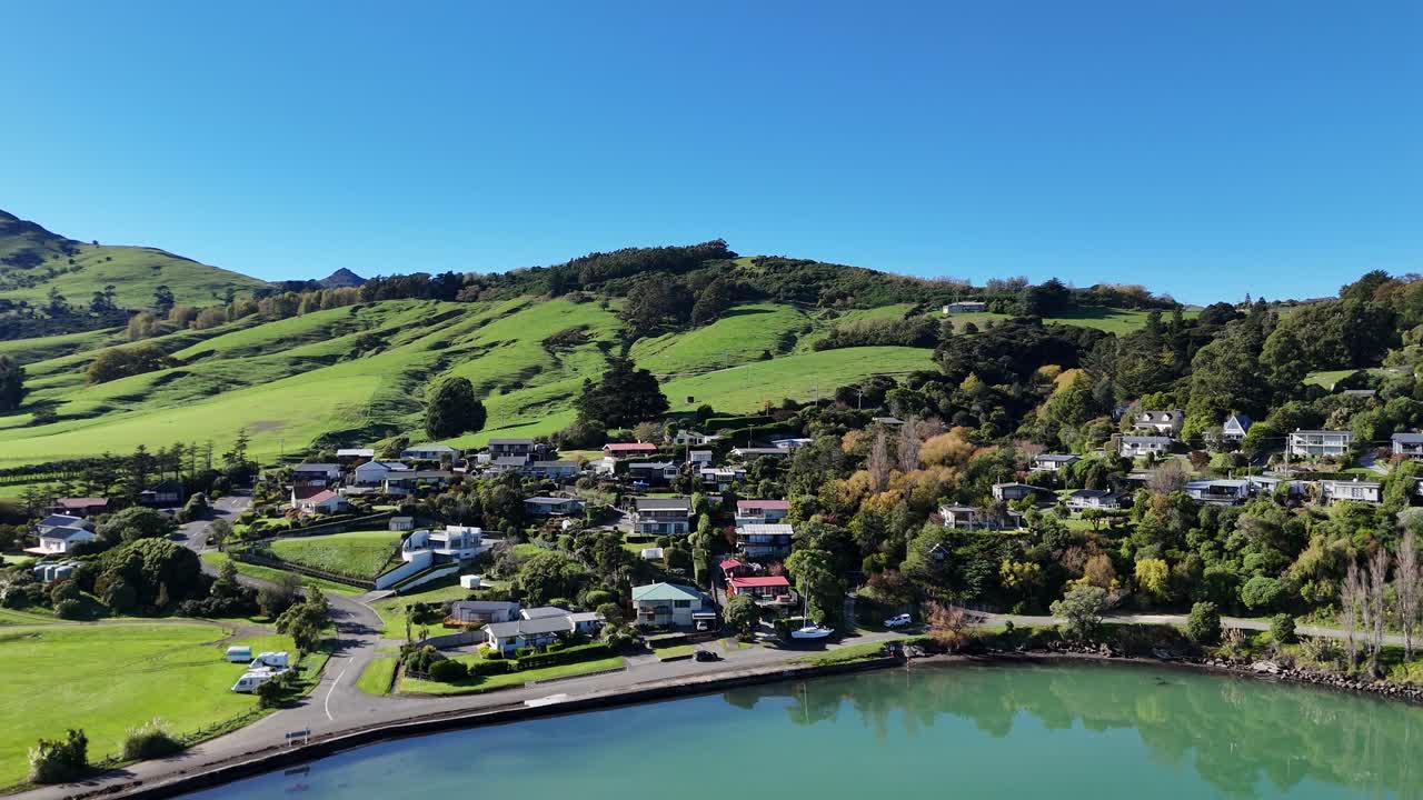 Drone footage captures Akaroa's lush hills, tranquil bay, and charming town under clear blue skies