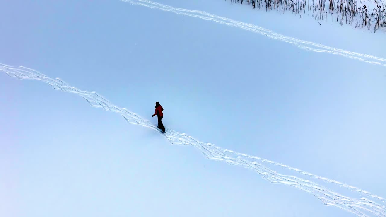 A person skis across a vast snow-covered field wearing a red jacket, creating fresh tracks through untouched terrain in a calm winter setting, viewed from an aerial perspective.