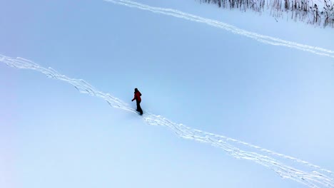 a person skis across a vast snow-covered field wearing a red jacket, creating fresh tracks through untouched terrain in a calm winter setting, viewed from an aerial perspective.