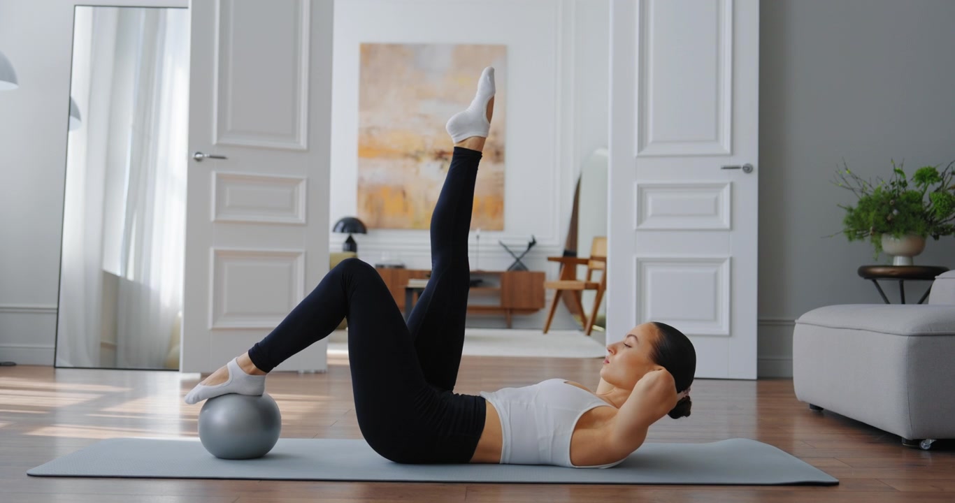 Giovane donna che esegue sollevamenti delle gambe Pilates con la palla da fitness a casa