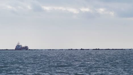 Large-red-cargo-ship-leaves-Port-of-Liepaja,-calm-bay-in-foreground,-wide-shot-from-a-distance