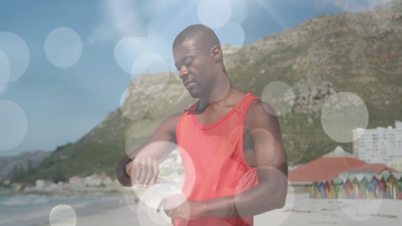 Animazione di luci brillanti su un uomo afroamericano che si allena in spiaggia usando uno smartwatch