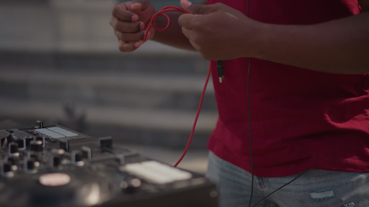 red bag revealing DJ cables outdoors, hands pull braided cable from torn red case, white sneakers and cobblestone plaza visible, connecting headphone lead to controller, focused preparation