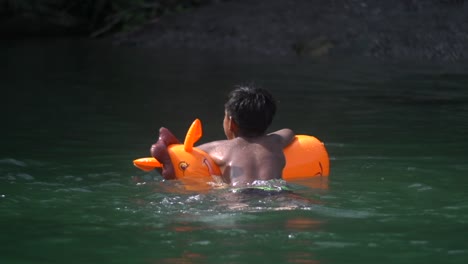 brown skinned indian child using orange inflatable boar to swim across river area filmed from backside