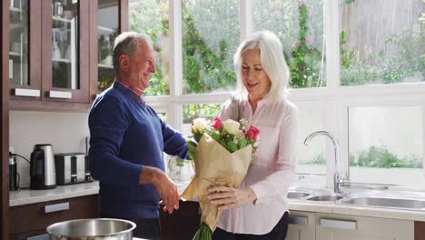 Un-Hombre-Mayor-Dándole-Un-Ramo-De-Flores-A-Su-Esposa-Y-Abrazándola-En-La-Cocina-De-Casa